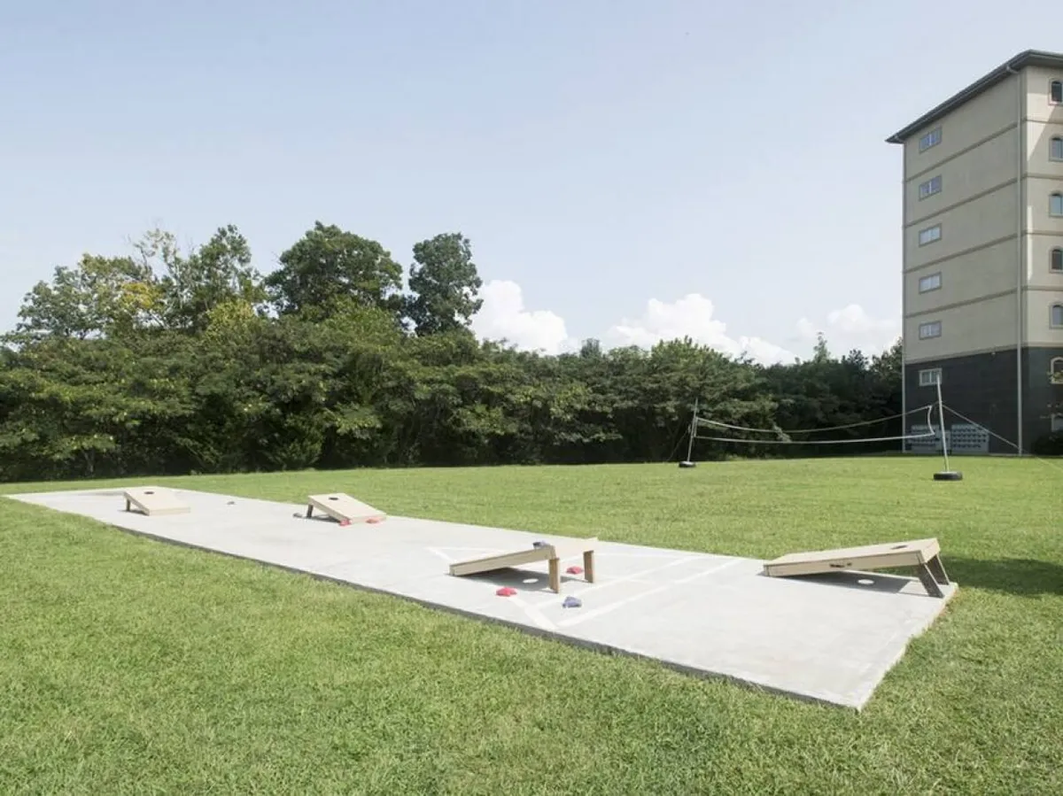 The image displays an inviting outdoor recreation area featuring cornhole boards and a volleyball setup on green grass, with a building nearby. The atmosphere is lively, highlighting opportunities for outdoor socializing and playful group activities.