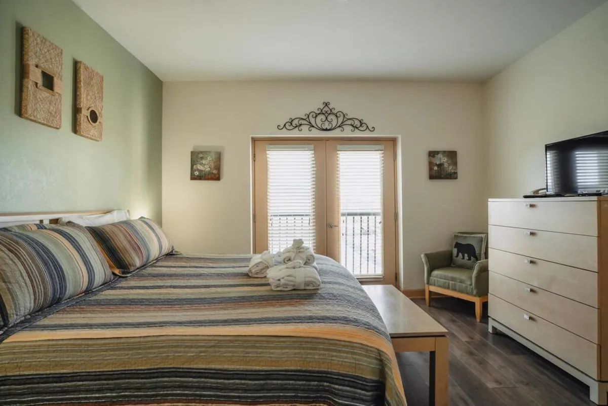 A serene bedroom with striped bedding, double doors opening to a private balcony, a dresser with a TV, and decorative wall art. The room feels calm and airy, bathed in natural light, with amenities that invite guests to relax after a busy day.