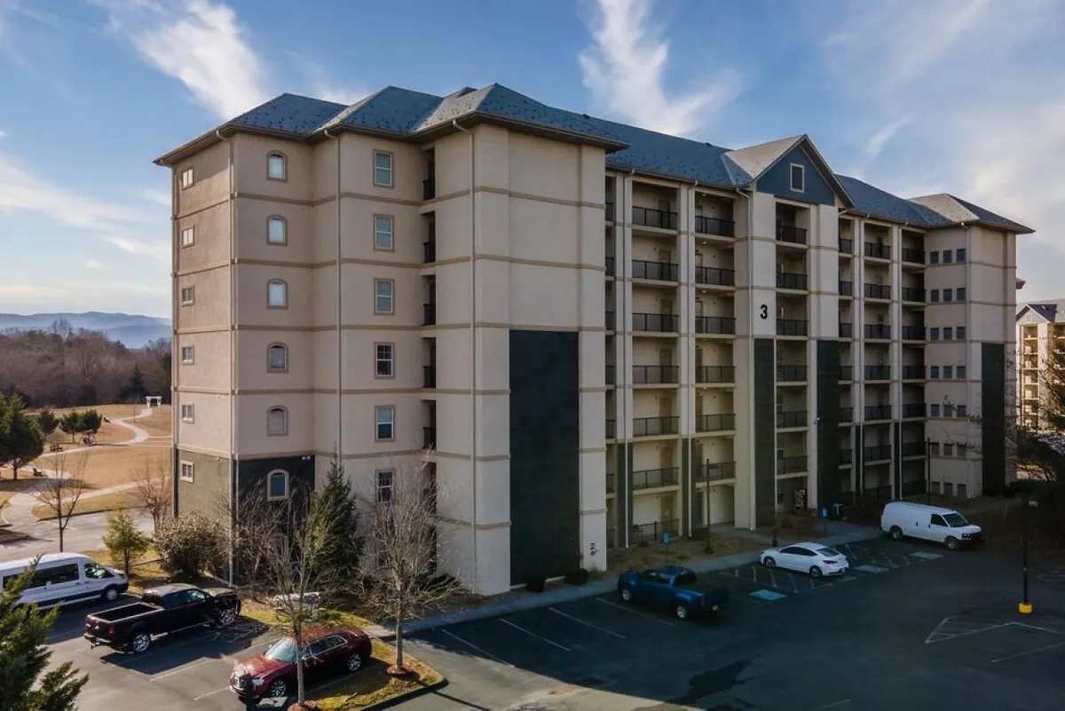 This exterior shot showcases the multi-story condo building surrounded by lush greenery and a spacious parking area. The landscape highlights the property's connection to nature and its inviting, contemporary design.