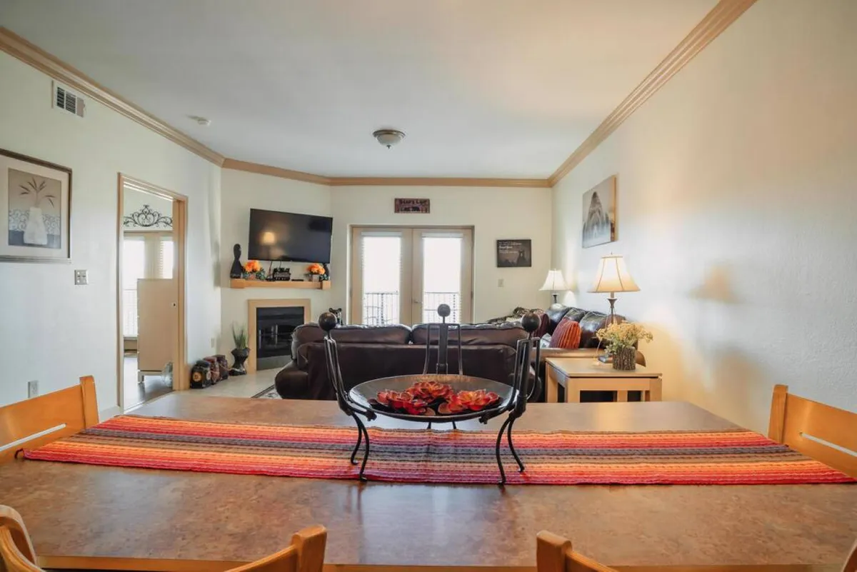 A bright and inviting living room with a comfortable sofa, a dining table adorned with a decorative bowl, and a flat-screen TV mounted on the wall. The space is softly lit with table lamps and features tasteful decor, creating a cozy atmosphere for relaxing and entertaining.