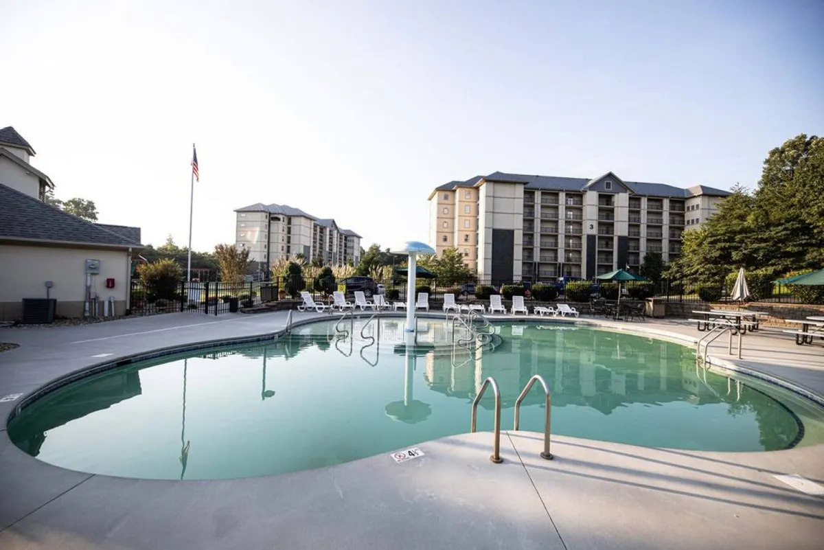 This image features a vibrant outdoor pool surrounded by loungers and lush trees, with resort buildings visible in the background. The pool area appears spacious and inviting, offering plenty of room for families to swim, sunbathe, and play together.