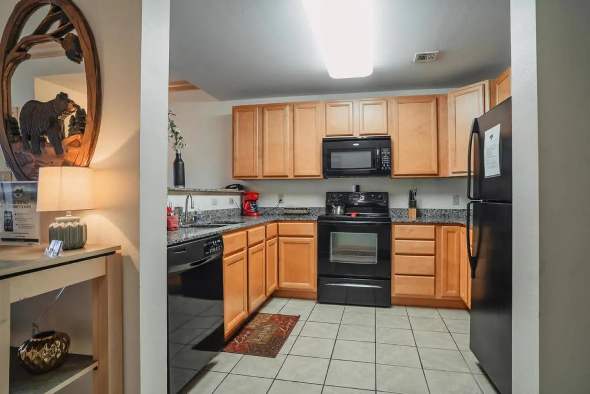 This image shows a well-equipped kitchen with sleek black appliances, granite countertops, warm wooden cabinets, a decorative lamp, and bear-themed artwork. The scene exudes a cozy and inviting atmosphere, suggesting a space that’s ready for both everyday meals and special family recipes.