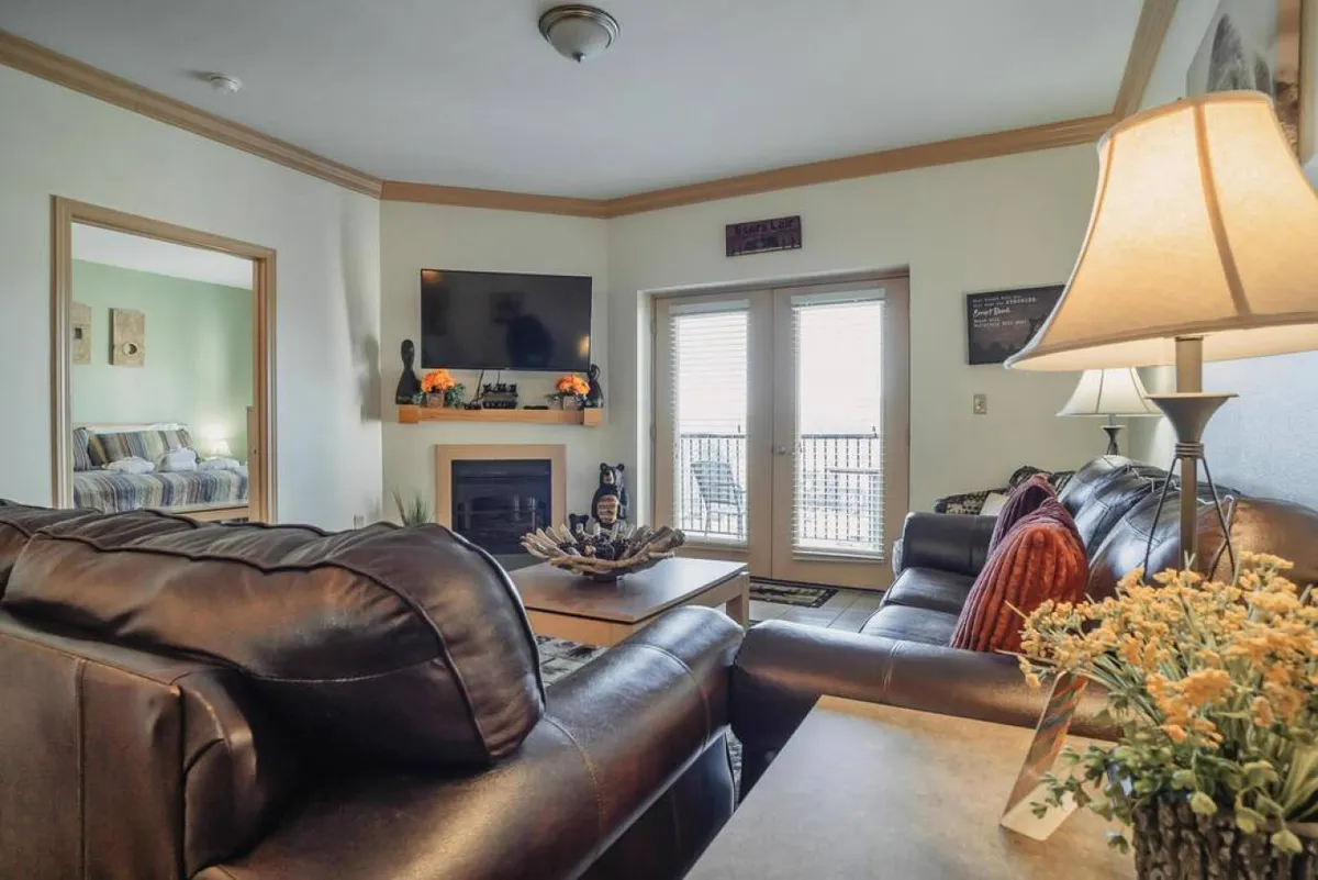 An interior scene reveals a stylish living area with leather sofas, a cozy fireplace, a TV, and sliding glass doors leading out to a balcony. The space is sunlit and inviting, creating a seamless connection between indoor relaxation and access to the outdoors.