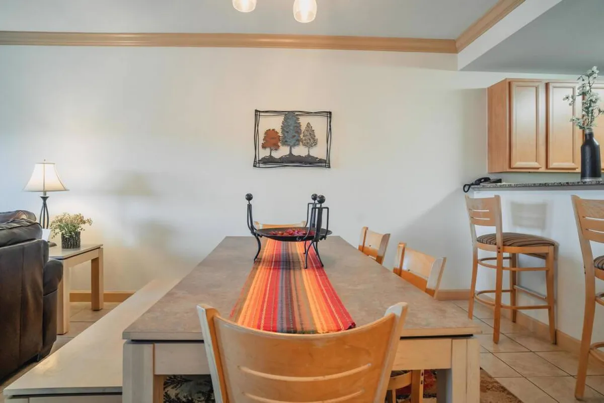 An inviting dining area with a spacious table, colorful table runner, lamp, framed artwork, and bar stools. The setup suggests a space made for family meals, festive celebrations, or quiet mornings with coffee.