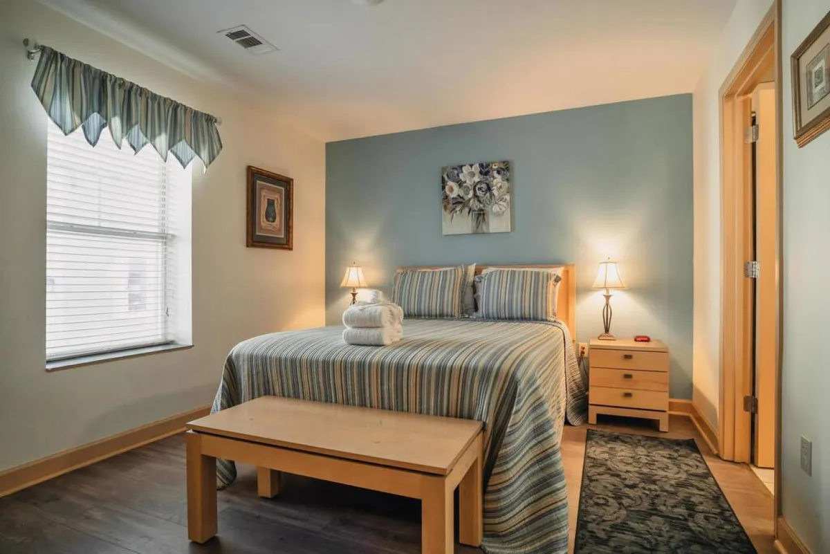 A bright, cozy bedroom featuring a plush bed topped with crisp linens, soft pillows, and inviting nightstand lamps. Artwork and a stylish bench at the foot of the bed add elegance, while natural light streams in, emphasizing the tranquil mood.