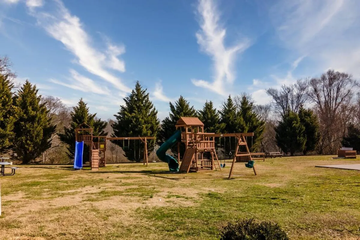 A lively playground scene with colorful equipment including swings and a slide, set on bright green grass and surrounded by trees. The setup is clearly designed for children, inviting exploration and active play.