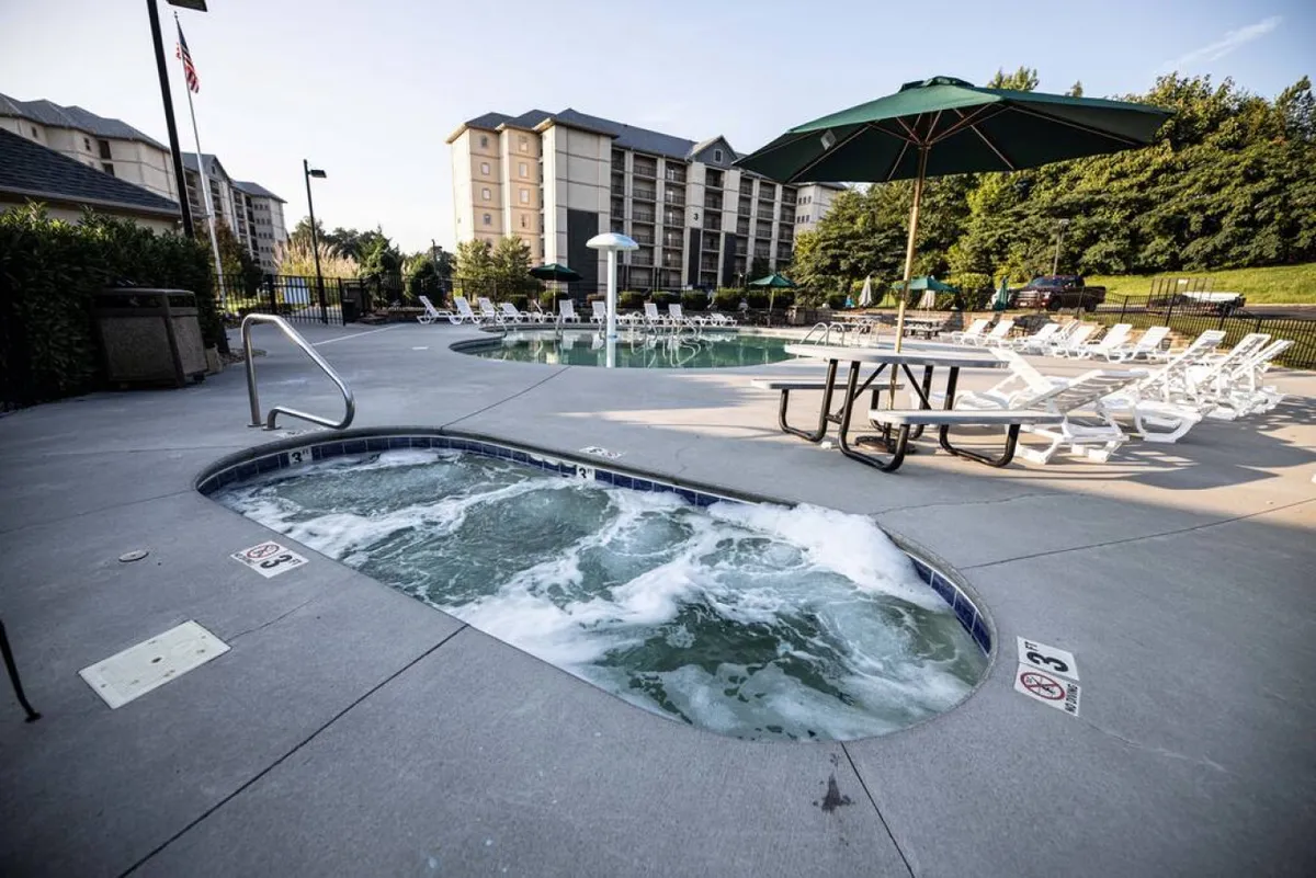 An outdoor pool area featuring a hot tub, swimming pool, lounge chairs, and a picnic table with umbrella. The space is ready for relaxation, group gatherings, and fun in the sun, surrounded by open green space.