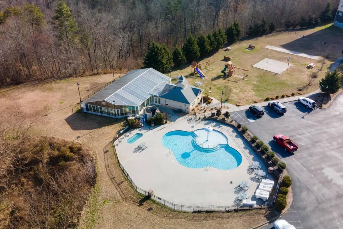 This joyful exterior shot shows an outdoor swimming pool adjacent to a recreational building and a playground area. Children can be seen playing, while families gather and relax by the pool in the sunshine.